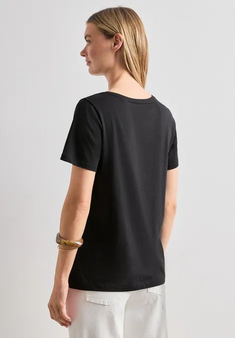 Frau von hinten in schwarzem T-Shirt und weißen Hosen, helle Haare über die Schulter fallend.