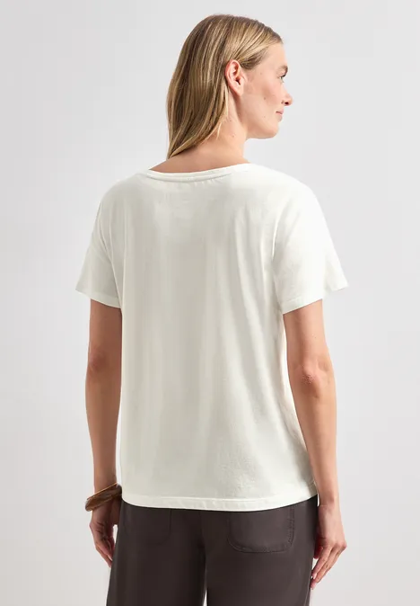 Eine Person steht mit dem Rücken zur Kamera, trägt ein weißes T-Shirt und eine dunkle, lockere Hose.