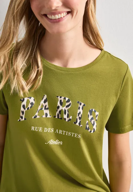 Eine Frau trägt ein olivgrünes T-Shirt mit Leopardenmuster-Schriftzug «PARIS RUE DES ARTISTES Atelier».