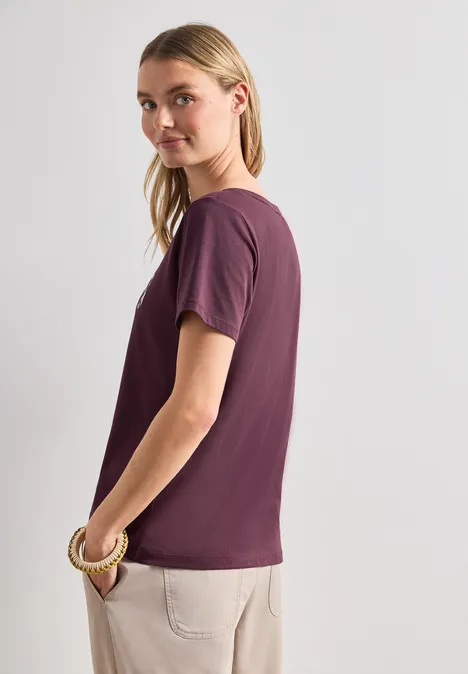 Seitlich stehende Person in lila T-Shirt und beigen Hosen, das Gesicht unkenntlich gemacht.