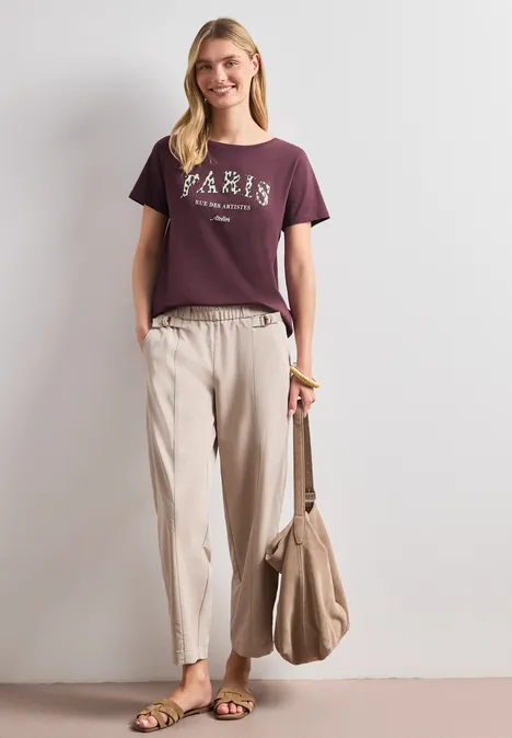 Frau mit burgunderfarbenem T-Shirt mit 'PARIS', beigefarbenen Hosen und Tasche an der Seite.