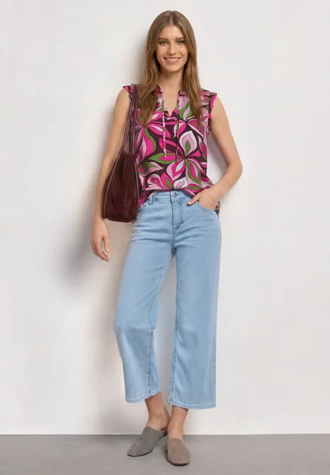 Frau steht vor hellem Hintergrund, trägt geblümte Bluse, hellblaue Cropped-Jeans, graue Loafers und braune Tasche.