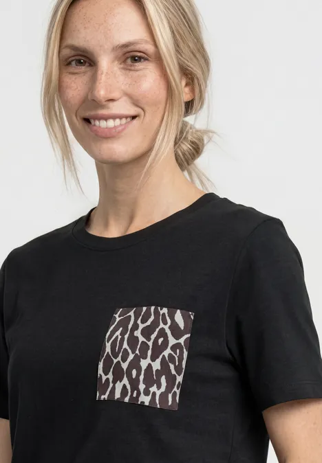 Une femme porte un t-shirt noir avec une poche à motif léopard sur fond clair.