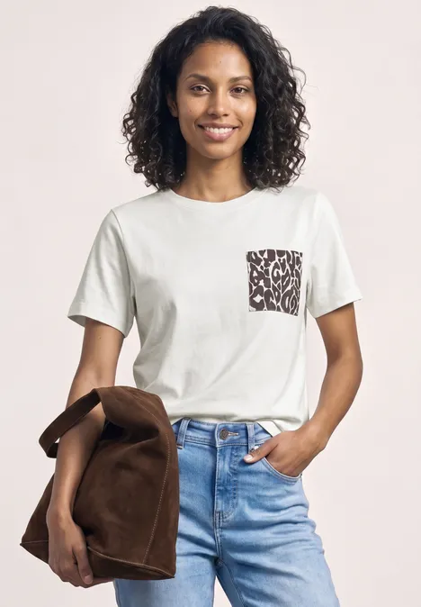 Iemand draagt een wit T-shirt met een patroonzak en een lichtblauwe spijkerbroek, terwijl hij een bruine jas vasthoudt.