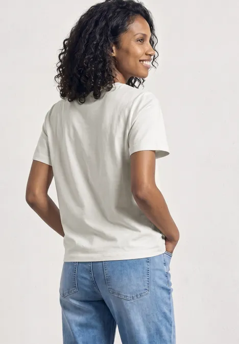Een persoon staat met de rug naar de camera, draagt een wit T-shirt en blauwe jeans, met donker krullend haar.