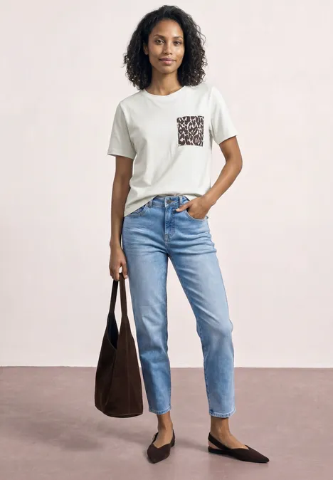 Vrouw in wit T-shirt met dessinzak, blauwe jeans, zwarte ballerina’s en grote bruine tas.
