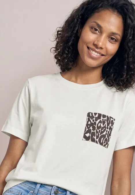 Een persoon draagt een wit t-shirt met een klein vierkant borstzakmotief en krullend haar.