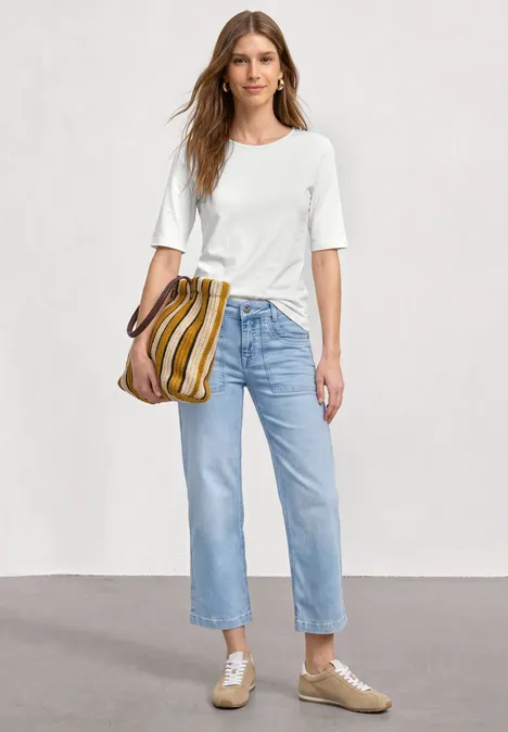 Eine Frau trägt ein weißes T-Shirt, hellblaue Jeans, beige Schuhe und eine gestreifte Tasche.