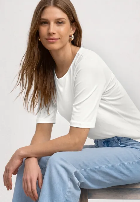 Eine Frau sitzt seitlich, trägt weißes T-Shirt mit Ellenbogen-Ärmeln und hellblaue Jeans.
