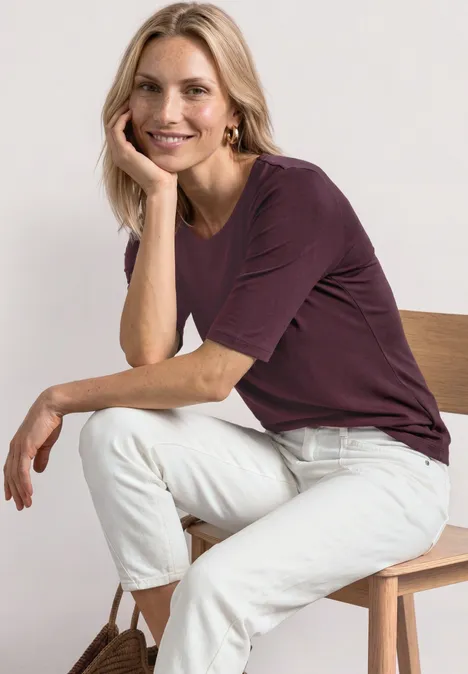 Frau mit blonden Haaren in lila Shirt und weißen Hosen sitzt entspannt auf einem Holzstuhl. Gesicht unscharf.