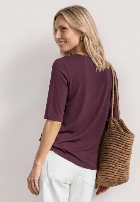 Dame mit lila T-Shirt, weißen Hosen und geflochtener Tasche blickt seitlich mit dem Rücken zur Kamera.