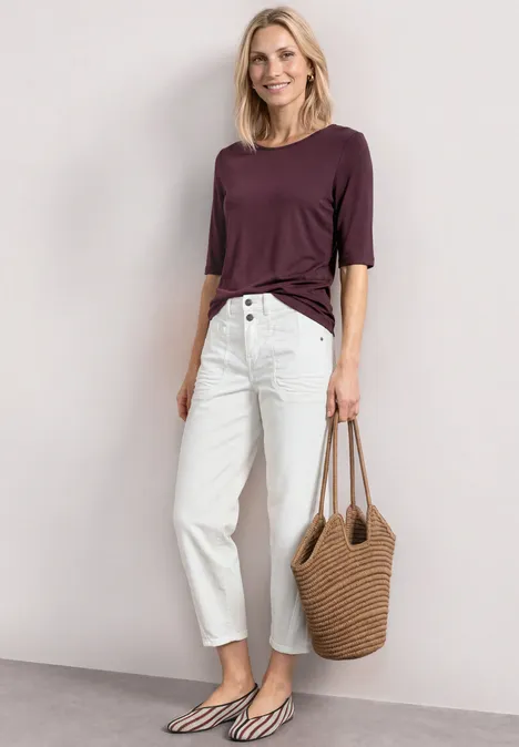 Frau in lila 3/4-Shirt, weißen High-Waist-Jeans, schwarz-weißen Ballerinas und Strohtasche.