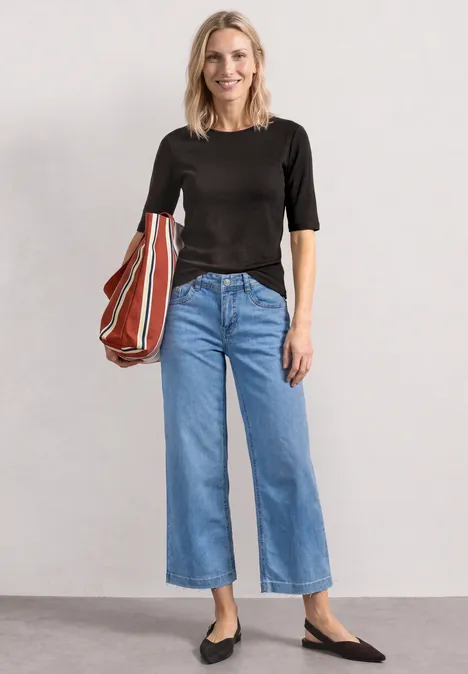 Frau trägt schwarze Kurzarmbluse, weite blaue Jeans, schwarze Ballerinas und hält eine gestreifte Tasche.