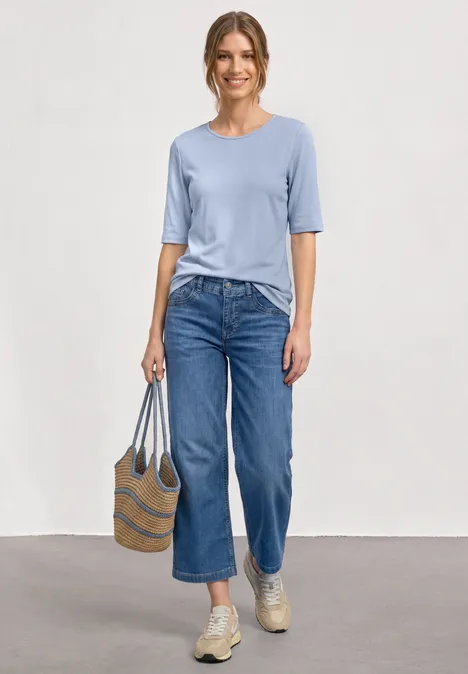 Eine Frau trägt ein hellblaues Shirt, weite, knöchellange Jeans, beige Sneaker und hält eine gestreifte Strohtasche.