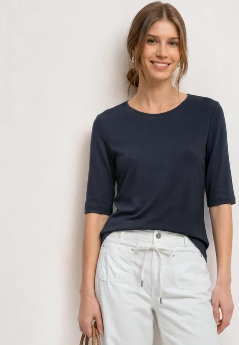 Frau trägt dunkelblaues Shirt und weiße Hose vor neutralem Hintergrund.