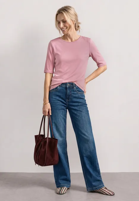 Eine Frau trägt ein rosafarbenes Shirt, blaue Jeans und hält eine braune Tasche.