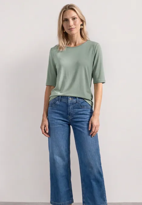 Frau in grünem T-Shirt und weiter Jeans steht vor neutralem Hintergrund.