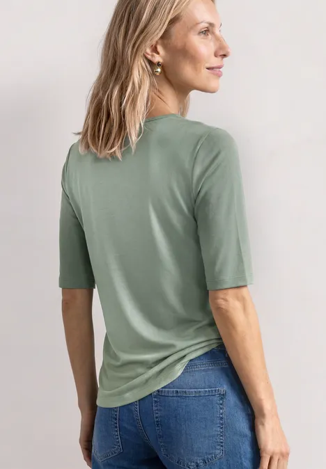 Frau von hinten in salbeigrünem Halbarmshirt und Jeans, helle Haare, dezente Ohrringe.