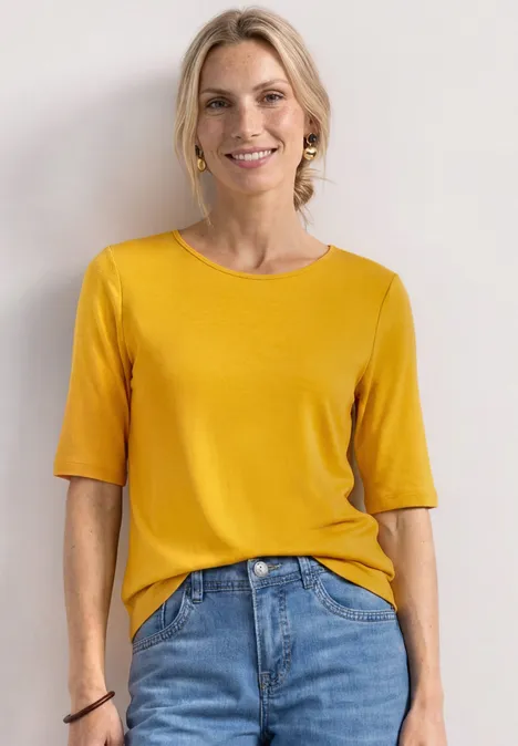Eine Frau mit gelbem T-Shirt und blauen Jeans steht vor heller Wand.