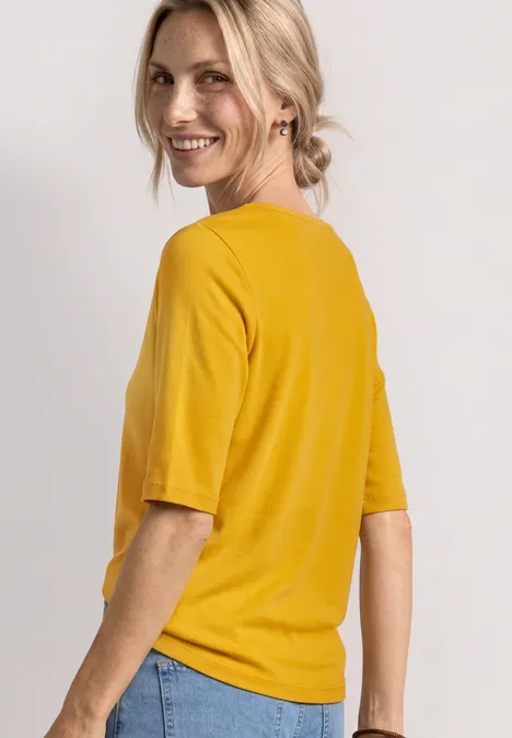Frau von hinten in gelbem T-Shirt und blauen Jeans vor hellem Hintergrund.