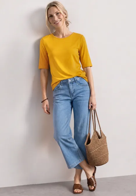 Frau lehnt an Wand, trägt gelbes T-Shirt, weite blaue Jeans, geflochtene Sandalen und Korbtasche.