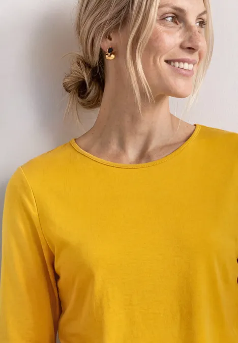 Person mit gelbem Oberteil, goldenen Creolen und Dutt vor hellem, neutralem Hintergrund.