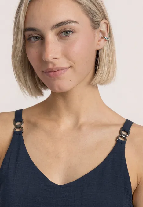 Junge Frau mit kurzem blondem Bob trägt dunkelblauen Top mit Riemen und Ohrschmuck vor hellem Hintergrund.