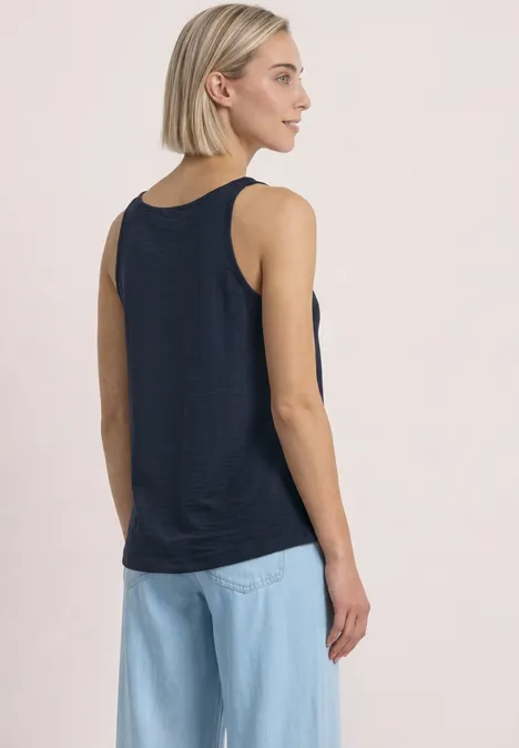 Frau von hinten in dunkelblauem Tanktop und hellblauen Jeans vor hellem Hintergrund.