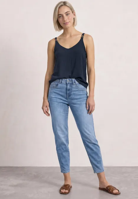 Eine junge Frau in schwarzem Top, blauen Jeans und braunen Sandalen vor neutralem Hintergrund.