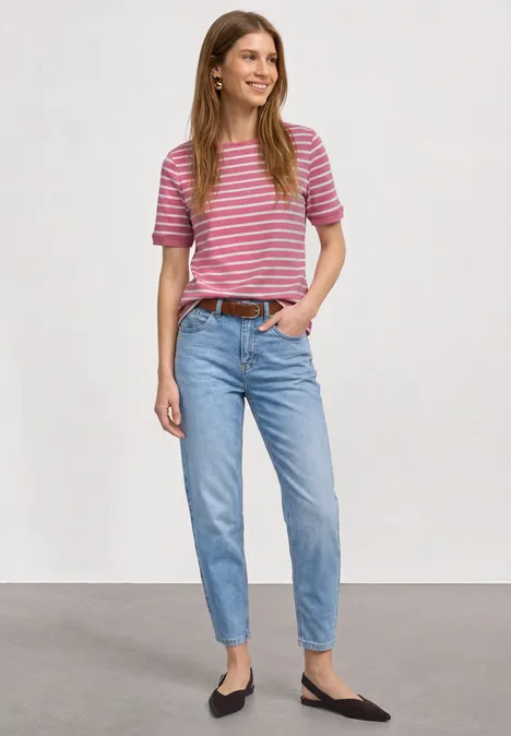 Eine Frau in rosa-weiß gestreiftem T-Shirt, blauen Jeans und schwarzen Ballerinas vor weißem Hintergrund.