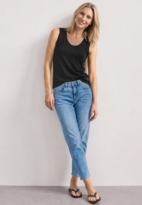 Frau in schwarzem Top, blauer Jeanshose und Flip-Flops steht lässig an einer hellen Wand.