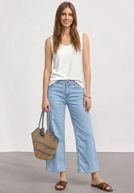 Femme en débardeur blanc, jean clair, sandales et sac en paille, debout devant un fond neutre.