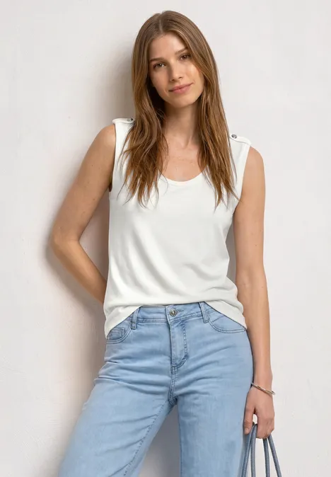 Une femme est debout devant un fond blanc, porte un haut blanc, un jean bleu clair et tient un téléphone.