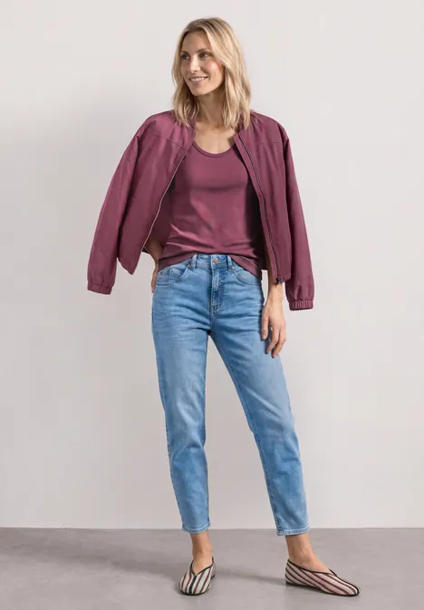 Femme portant une veste violette, un t-shirt bordeaux, un jean bleu clair et des tongs rayées sur fond neutre.