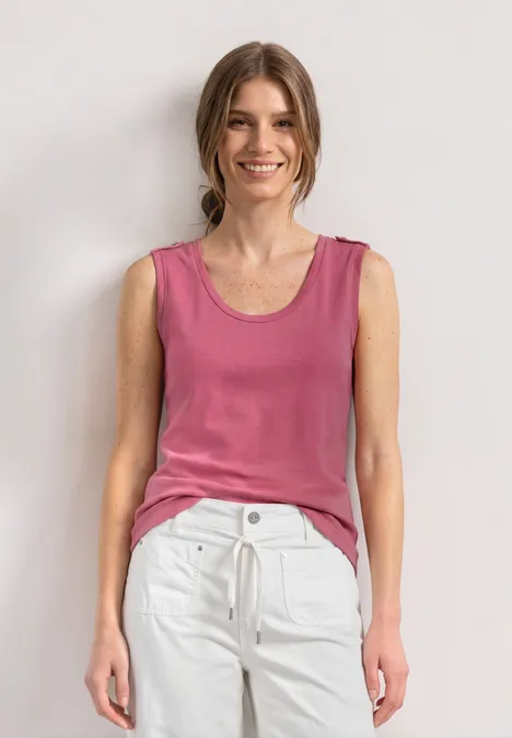 Haut à col V arrondi avec boutons aux épaules blush berry Eine Frau steht vor weißem Hintergrund, trägt rosa Top und weiße Hose.