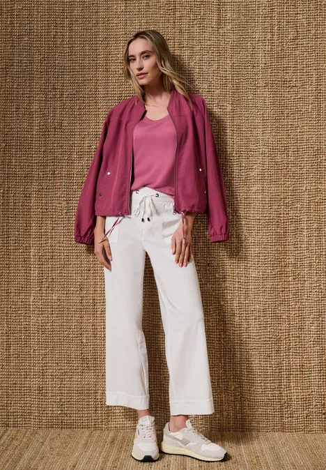 Haut à col V arrondi avec boutons aux épaules blush berry Frau trägt pinke Jacke und Top, weiße weite Hose, steht vor strukturierter Wand.