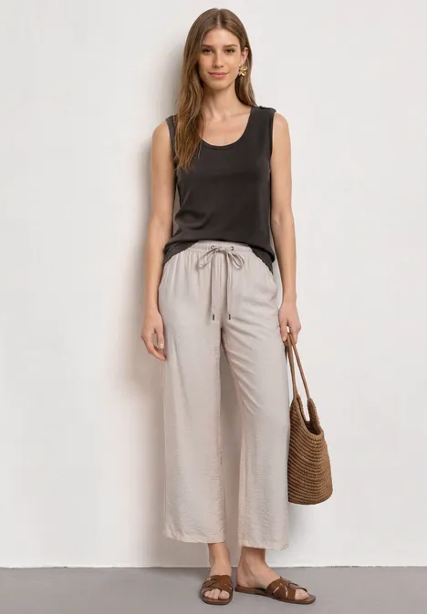 Haut à col en V et boutons black coffee Frau trägt braunes ärmelloses Top, helle, weite Leinenhose, Basttasche und flache Sandalen.