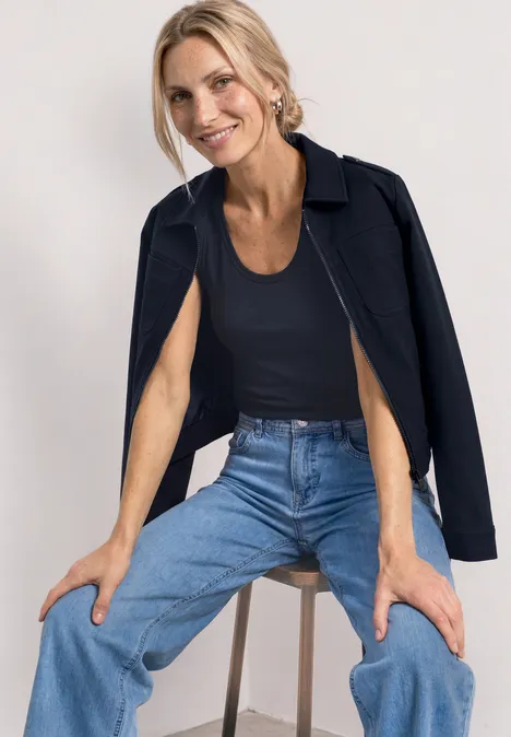 Haut à col V arrondi avec boutons aux épaules darkest blue Frau sitzt auf Hocker, trägt blaue Jeans, schwarzes Top und Jacke über den Schultern.