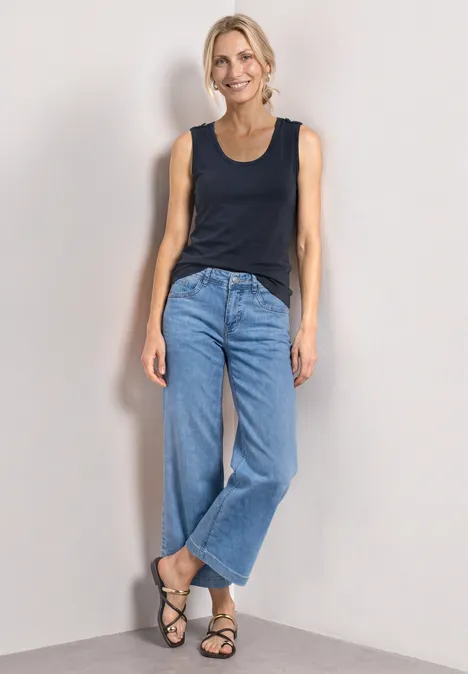 Haut à col V arrondi avec boutons aux épaules darkest blue Eine Person in schwarzem Top, weiten Jeans und Sandalen lehnt lässig vor heller Wand.