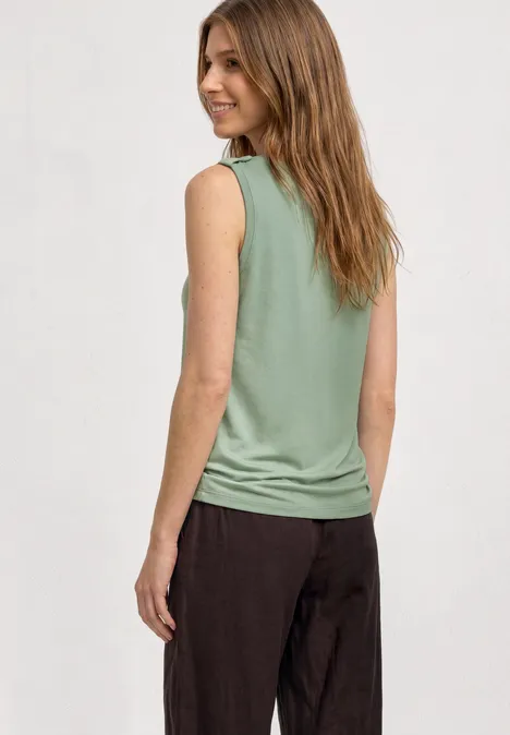 Top mit V-Neck und Knopfdetail eucalyptus green Frau von hinten in grünem ärmellosen Shirt und dunklen Hosen vor hellgrauem Hintergrund.