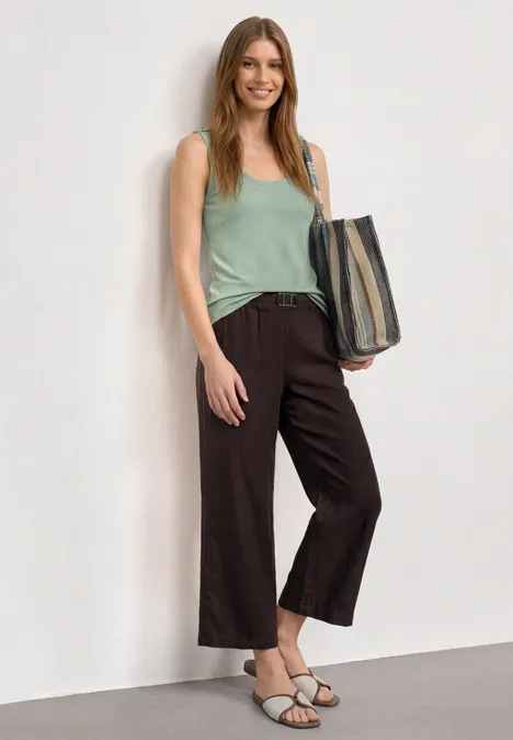 Top mit V-Neck und Knopfdetail eucalyptus green Frau in grünem Top und weiten, dunklen Hosen mit Tasche und Sandalen vor hellem Hintergrund.