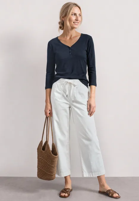 Frau in blauem Shirt, weißen weiten Hosen, braunen Sandalen und Strohtasche vor neutralem Hintergrund.