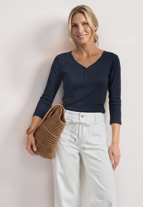 Frau in dunkelblauem Langarmshirt und weißen Hosen mit Strohtasche vor hellem Hintergrund.