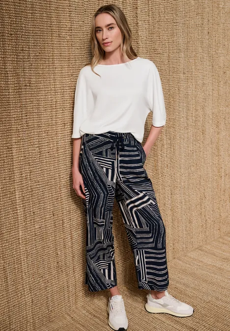Basic Shirt met boothals en elastische zoom off white Vrouw voor geweven achtergrond met witte top, wijde patroonbroek en witte sneakers.