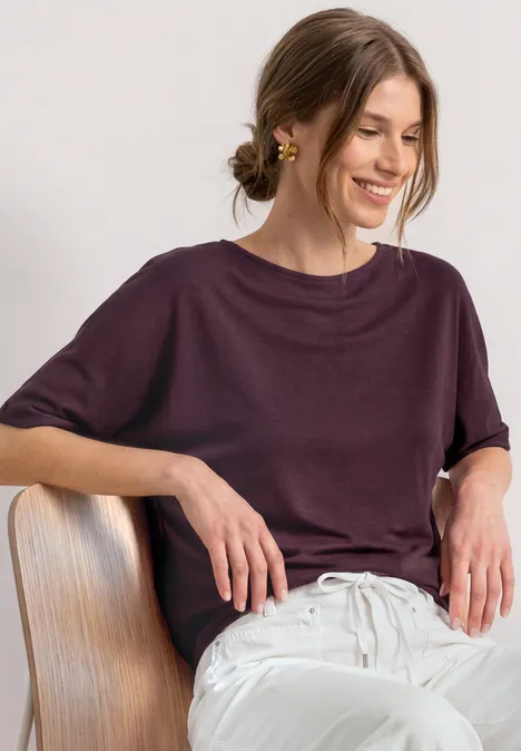Basic Shirt met boothals en elastische zoom rustic rouge Een glimlachende vrouw zit op een stoel, draagt een losvallende paarse top en een witte broek.