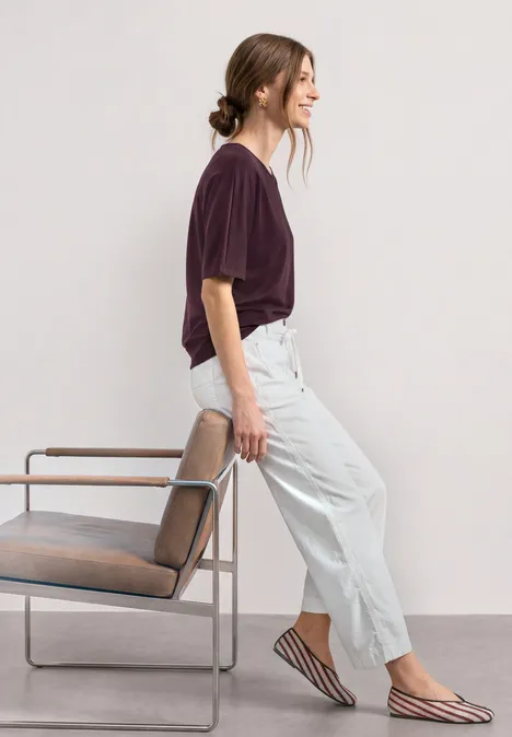 Basic Shirt met boothals en elastische zoom rustic rouge Vrouw in paarse T-shirt, lichte broek en gestreepte ballerina's leunt tegen een stoel voor een lichte achtergrond.