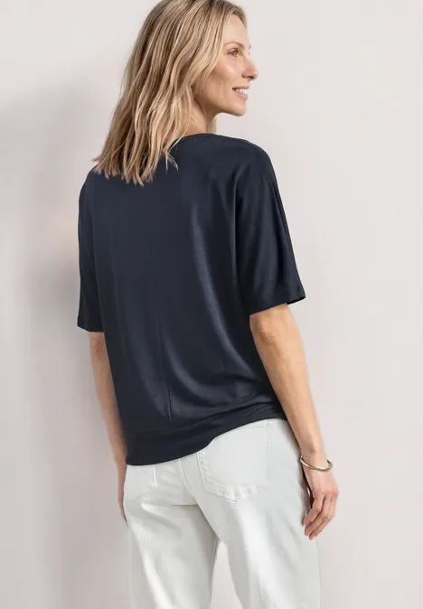 Eine Person steht seitlich mit Blick auf den Rücken, trägt ein dunkelblaues T-Shirt und weiße Jeans.