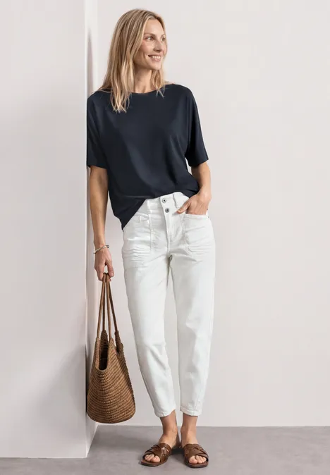 Frau lehnt an Wand, trägt schwarzes T-Shirt, weiße Hose und geflochtene Tasche.