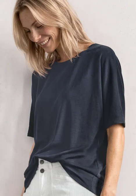 Eine Frau in Navy-T-Shirt und weißen Hosen lächelt und blickt nach unten vor hellem Hintergrund.