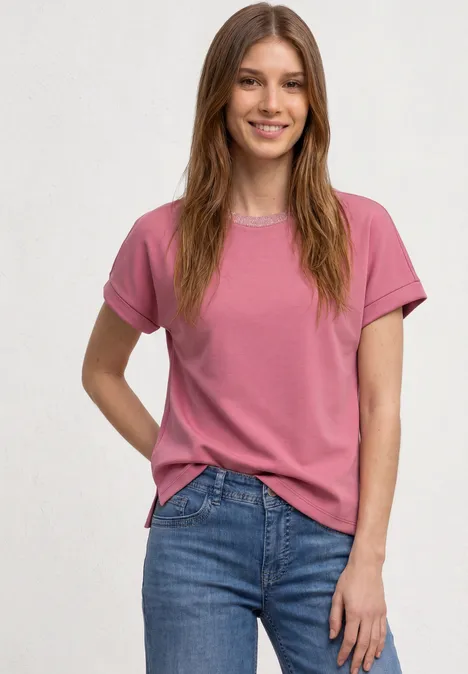 Eine lächelnde Frau steht vor hellem Hintergrund, trägt ein rosa T-Shirt und blaue Jeans.